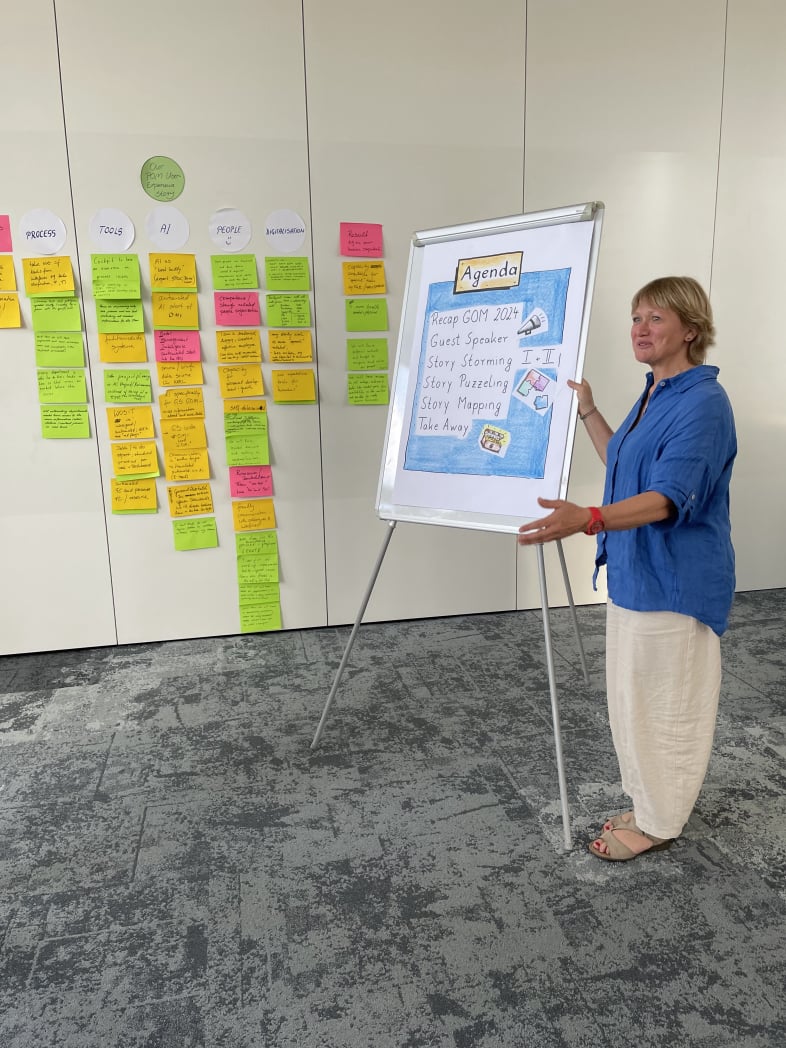 Marion Thomas steht mit mit mittelblauer Kurzarmbluse und beger Hose neben einem Flipchart.