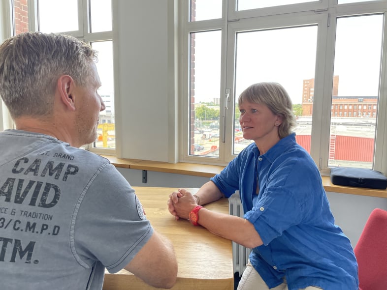 Marion Thomas mit kurzen blonden Haaren und knallblauer kurzärmlicher Bluse sitzt an einem Tisch, die Arme darauf abgelegt. Sie sitzt rechts und schaut zu einem Mann, mit dem sie sich unterhält. Sie schaut freundlich und aufmerksam den Mann an.